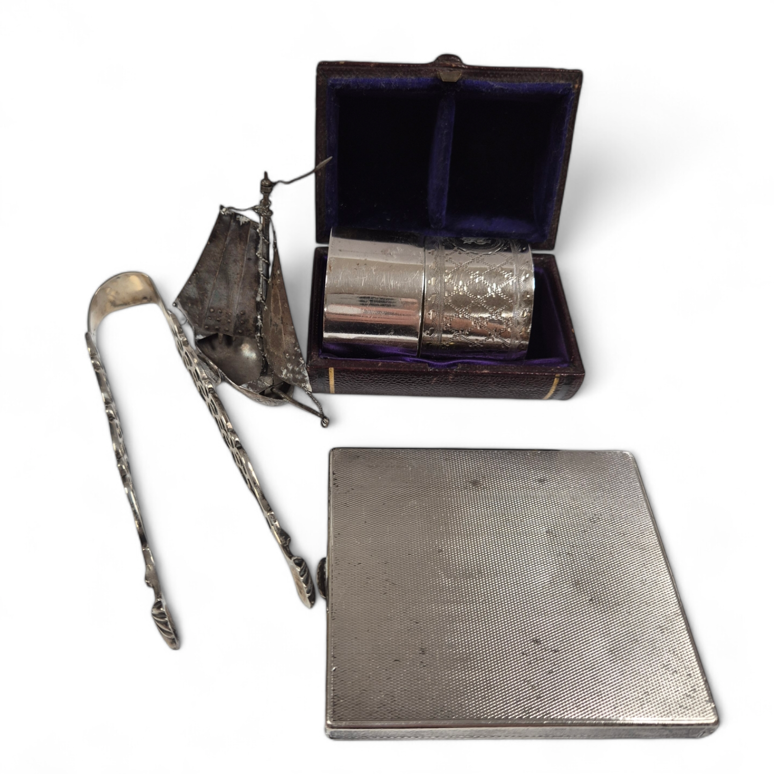 A 1930's engine turned silver cigarette case, 88mm, two silver napkin rings, a white metal miniature model of a boat and a pair of Georgian silver sugar tongs (a.f.). Condition - poor to fair